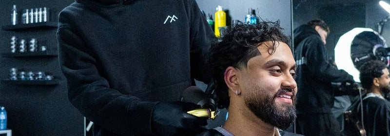 Barber at Montana Fades in Oakville, Ontario wearing a black branded hoodie and gloves using a gold clipper to shape a client’s fade haircut as the client smiles in a modern shop setting.