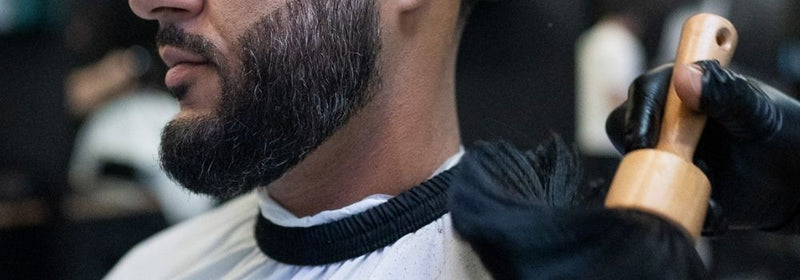 Barber at Montana Fades in Oakville, Ontario brushing loose hair clippings off a client’s trimmed beard with a wooden-handled brush after a precision fade.